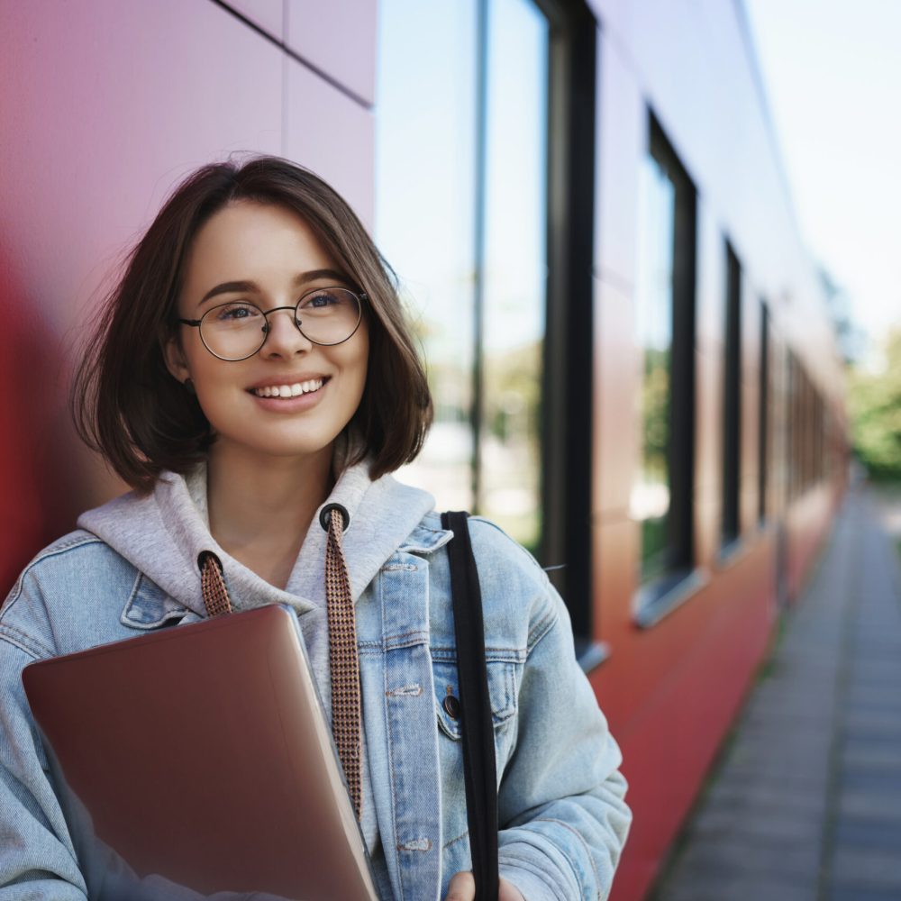 Vertical portrait of happy dreamy young female student look away with pleased expression, smiling, holding laptop on her way back to dormitory or work, enjoying spring sunny day, study with friend.